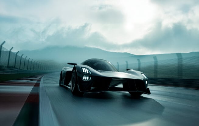 A sleek, futuristic car speeds on a race track under a cloudy sky. The vehicle's design features sharp angles and bright headlights. The surrounding environment appears wet, with mountains visible in the background.