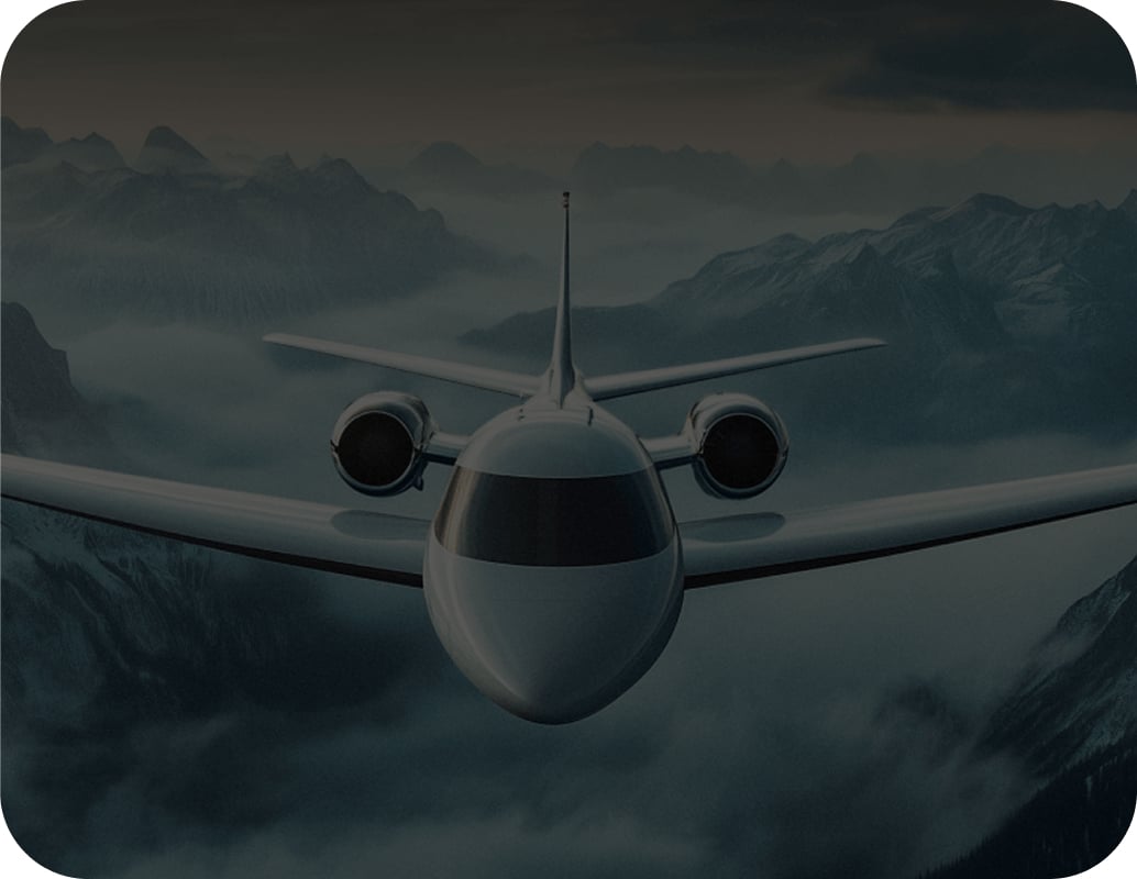 A jet airplane flies above misty mountains at dusk, with dramatic clouds and peaks in the background. The aircraft is shown head-on, dominating the center of the image.