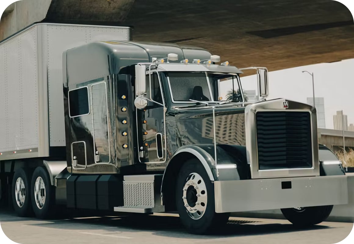 A shiny black semi-truck with chrome details and a large front grille is parked under an overpass in an urban area, with city buildings visible in the background.