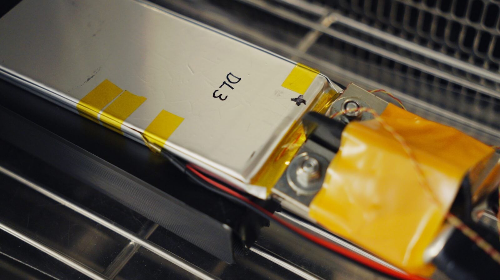 Close-up of a rectangular battery cell secured with yellow tape, labeled "D13," connected by wires, sitting on a metal rack.