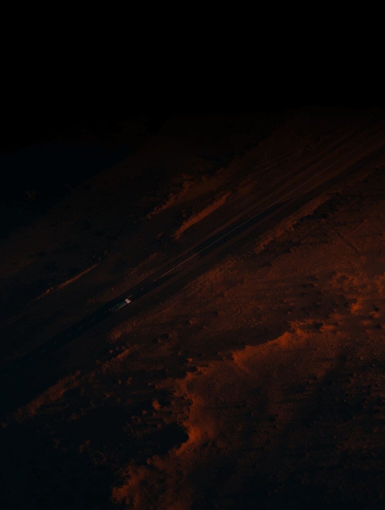 Aerial view of a car traveling on a long, straight road cutting through a desert landscape. The surroundings have a dark, earthy tone with sparse vegetation, and the road appears dimly lit under the evening sky.