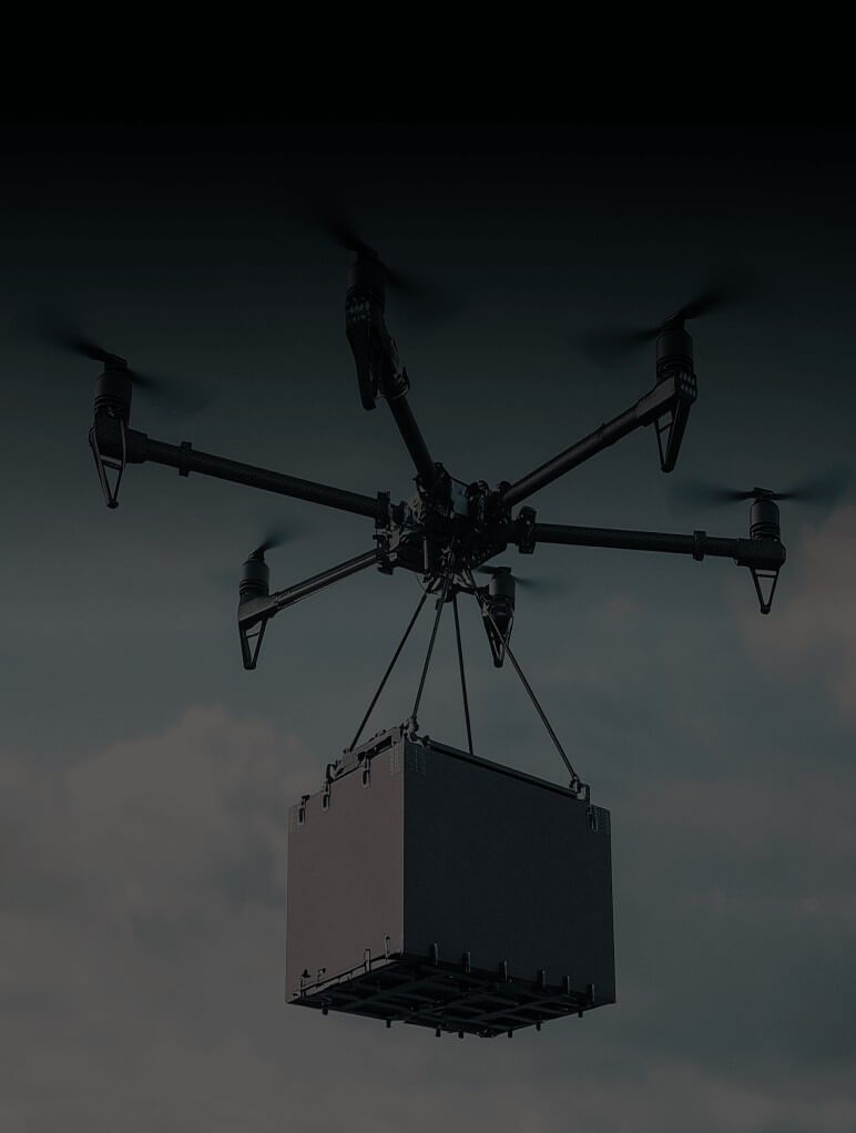 A drone with six rotors is carrying a large, rectangular package suspended below it against a cloudy sky.